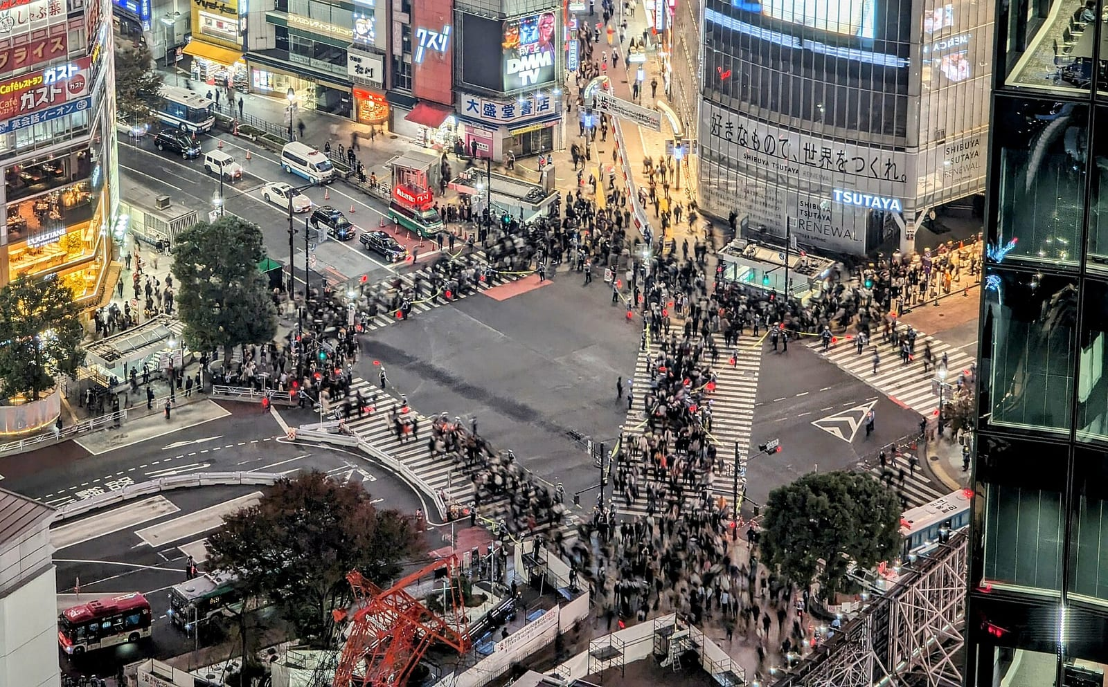 Yüksek bir binadan çekilmiş, Shibuya'daki ünlü yaya geçidinde kalabalığın dört yöne doğru karşıdan karşıya geçtiği akşam manzarası ve çevredeki binalar.