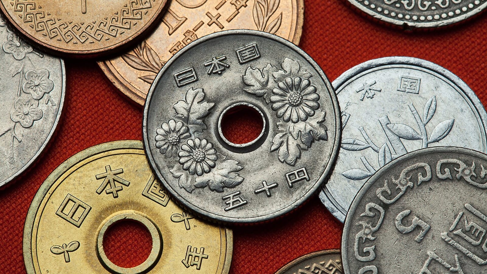 Kırmızı bir zemin üzerinde, ortasında delik bulunan 50 yenlik madeni para ve çevresinde farklı değerlerde birçok Japon bozuk parası yakından görünüyor.