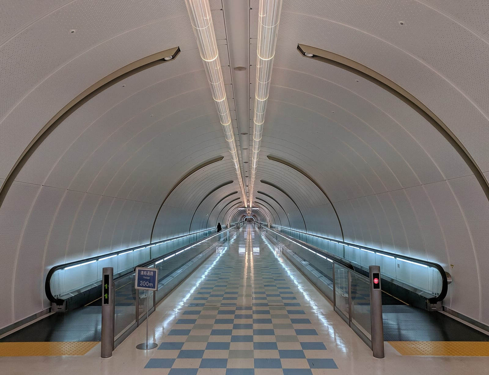 Narita Havalimanı 1. Terminal'de iki yanında yürüyen bant bulunan, aydınlık ve kemerli uzun bir yeraltı geçidi ile ortasında damalı zemin görünüyor.