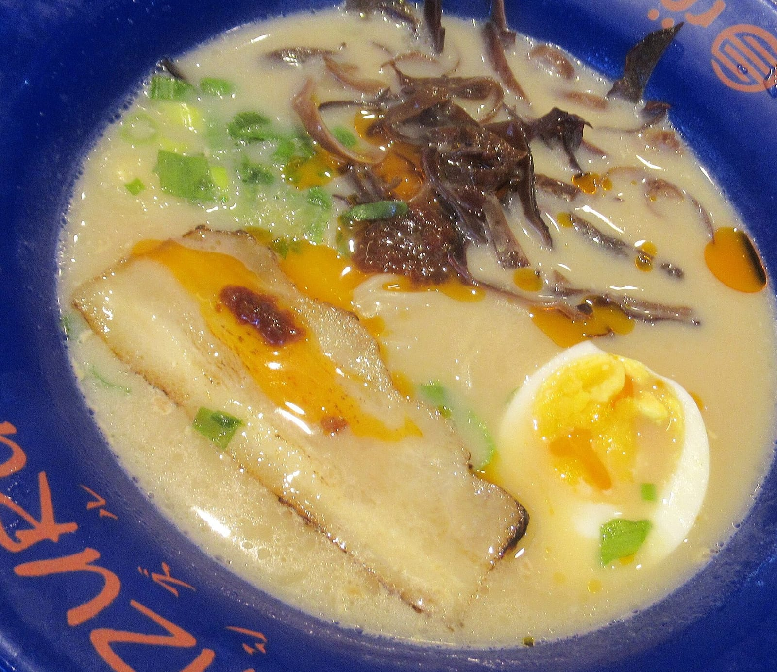 Mavi bir kasede krema renkli tonkotsu ramen çorbası, üzerinde domuz eti dilimi, yumurta, mantar ve taze soğan bulunuyor.