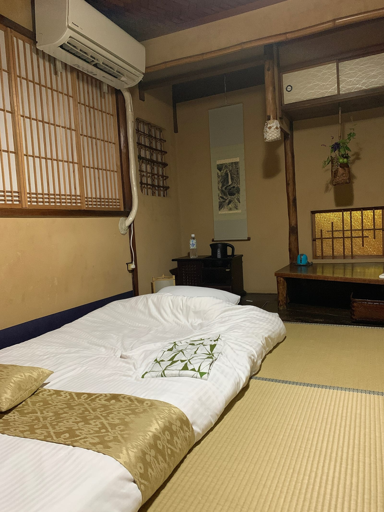 Geleneksel bir ryokan odasında tatami zemin üzerine serilmiş beyaz futon, şoji pencereler, asılı resim ve alçak ahşap mobilyalar görünüyor.