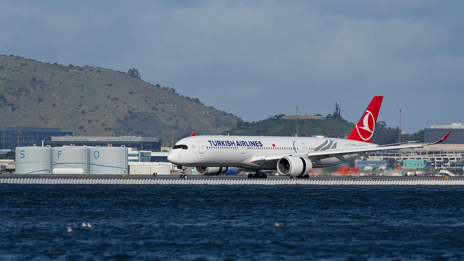Kırmızı kuyruklu beyaz bir Turkish Airlines Airbus A350 uçağı, arkasında yeşil tepeler olan deniz kıyısındaki pistte ilerliyor.