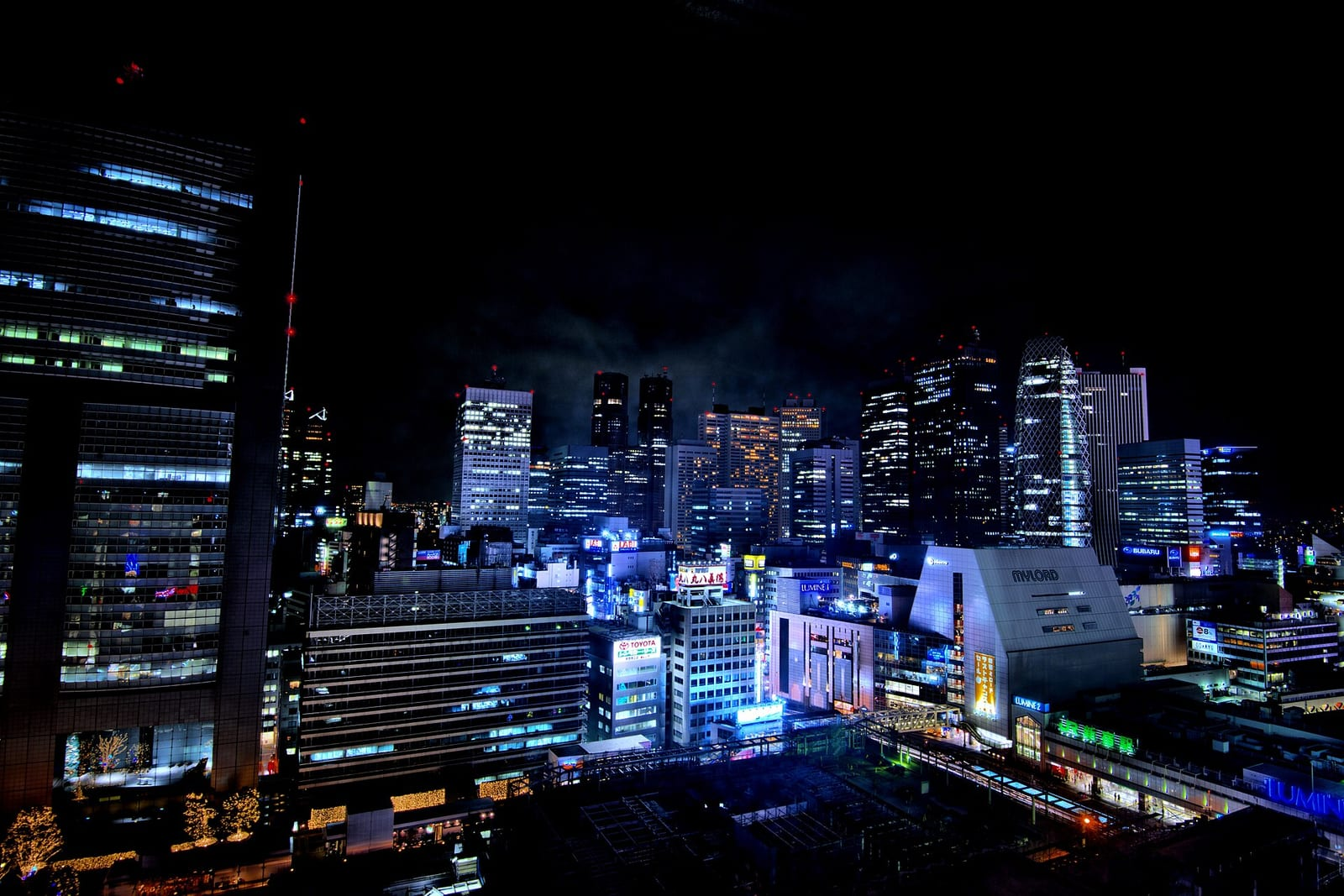 Shinjuku istasyonu çevresinde geceleyin ışıklı gökdelenler ve mavi tonlu şehir manzarası yukarıdan çekilmiş şekilde görülüyor.