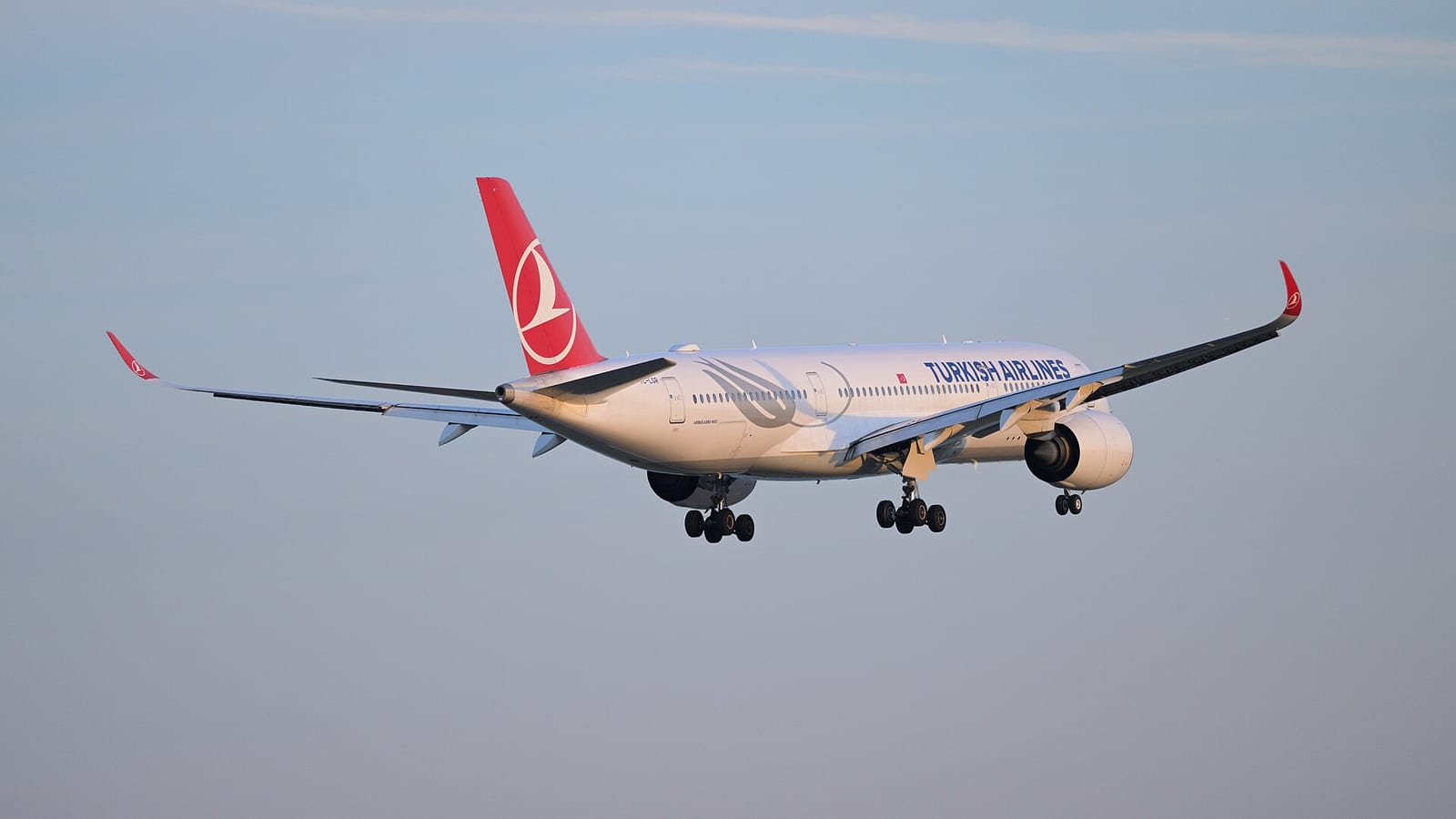 Türk Hava Yolları'nın kırmızı-beyaz Airbus A350 uçağı, açık mavi gökyüzünde inişe geçerken iniş takımları açık halde havada görülüyor.