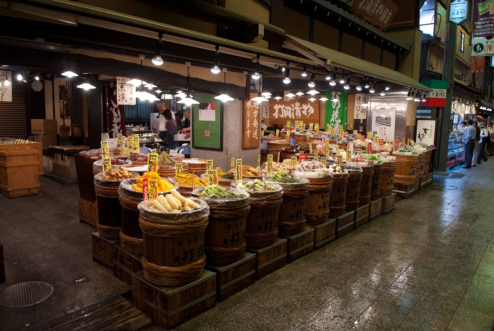 Kyoto Nishiki Pazarı'nda ahşap fıçılarda turşu ve sebzeler satan bir dükkan, loş sokakta ışıklarla aydınlatılmış halde görülüyor.