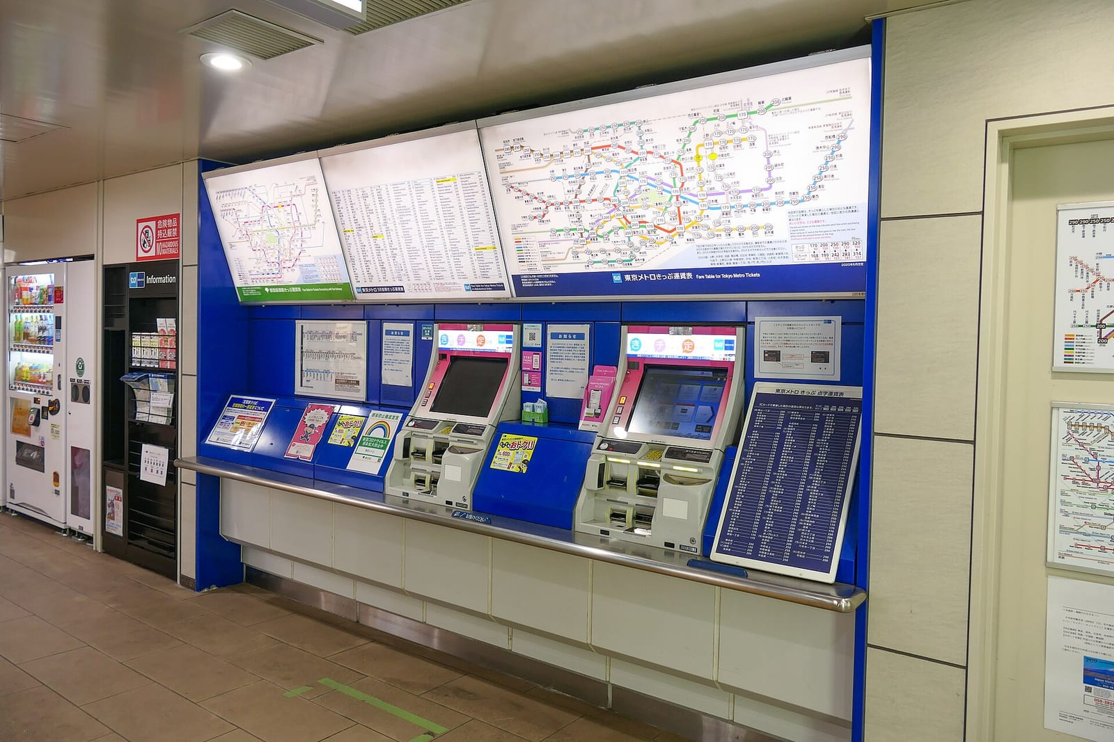Tokyo Metro istasyonunda mavi renkli bilet otomatları ile üstlerinde asılı renkli hat haritası ve ücret tablosu net biçimde görülüyor.