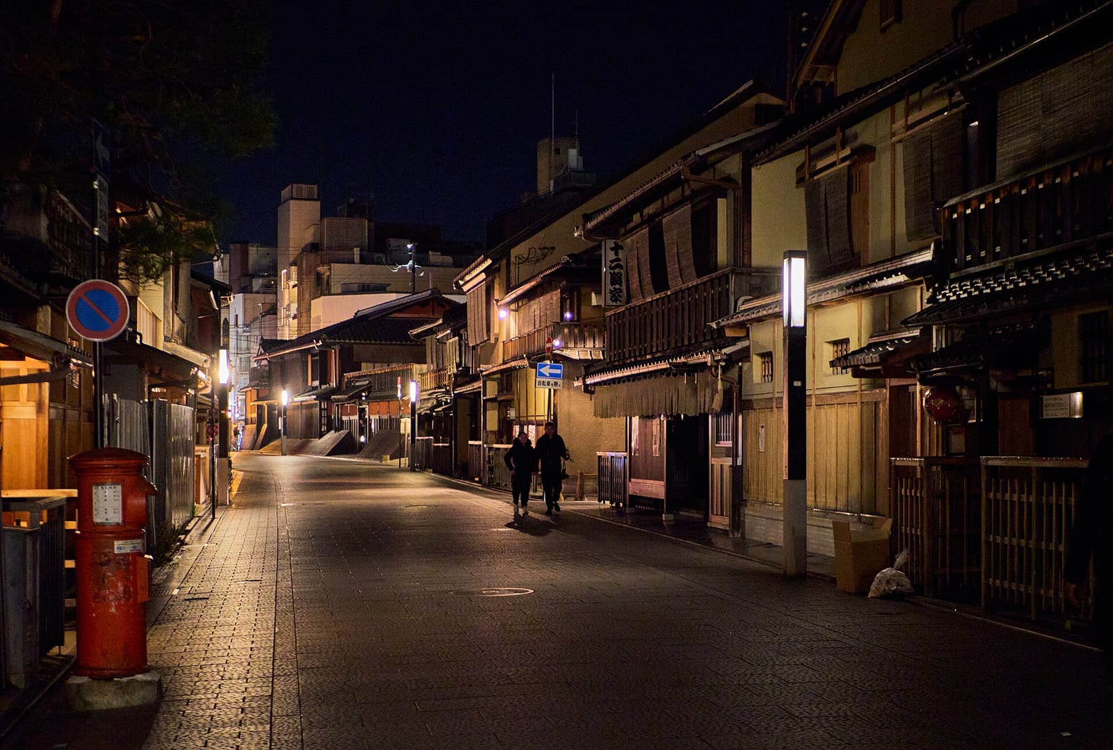 Kyoto'nun Gion semtinde gece, geleneksel ahşap machiya evleri arasındaki sakin sokakta yürüyen iki kişi ve kırmızı bir posta kutusu görülüyor.