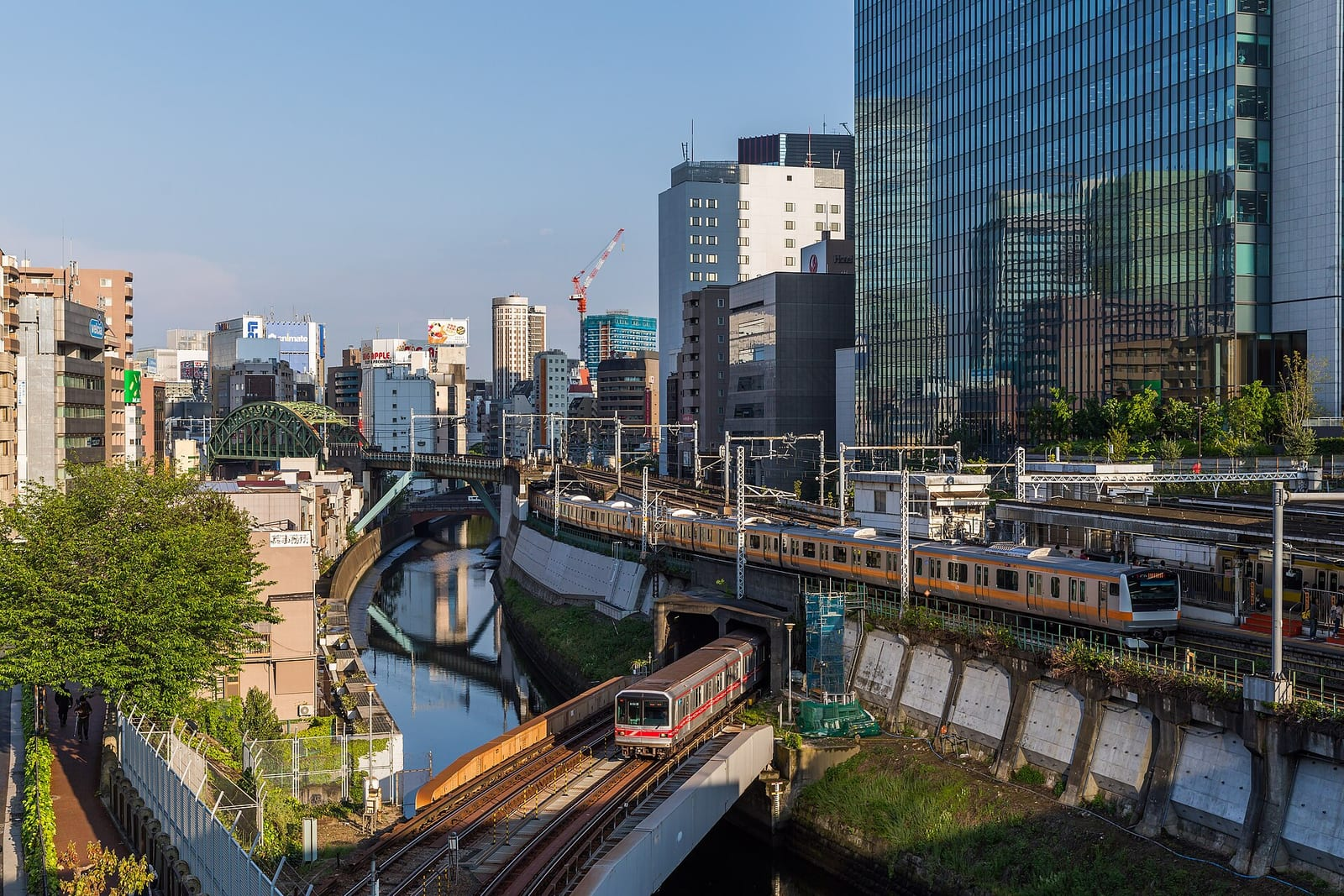 Tokyo'da bir nehir kıyısında, yüksek binaların arasından geçen kırmızı metro ile turuncu çizgili banliyö treni ve altlarındaki köprüler görülüyor.