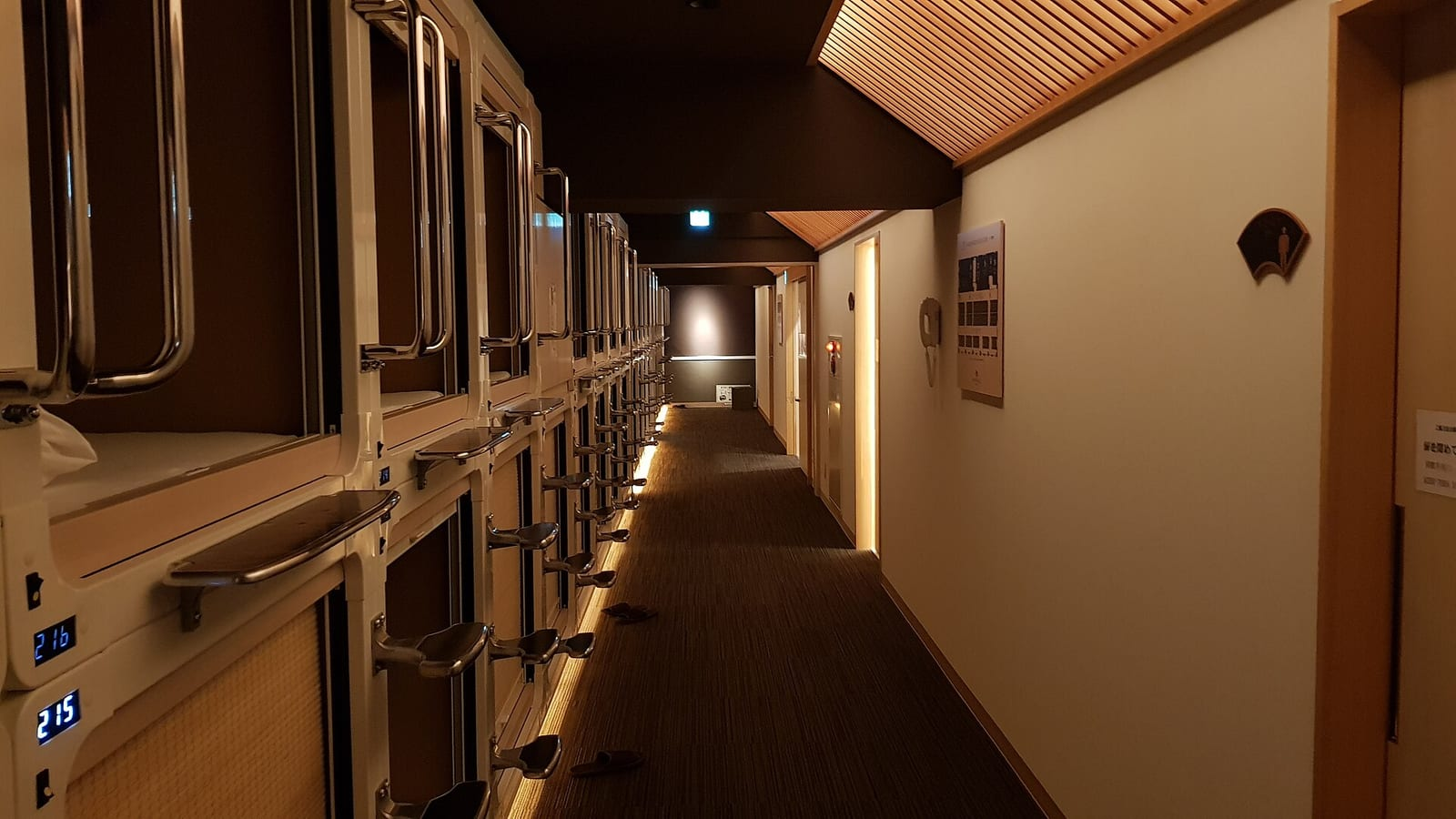 Tokyo'da bir kapsül otelde loş ışıklı koridor boyunca iki yanda dizilmiş kapsül yatak üniteleri ve ahşap tavan detayları.