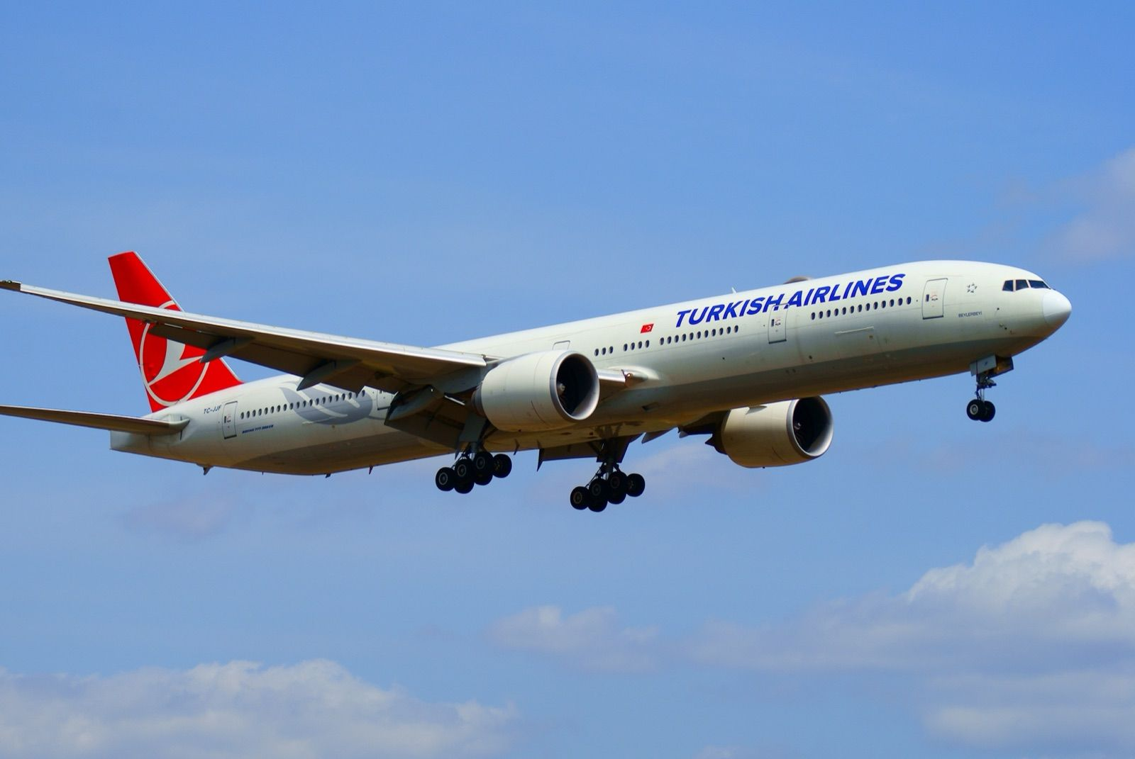 Kırmızı kuyruklu beyaz bir Turkish Airlines Boeing 777-300ER uçağı, mavi gökyüzünde iniş takımları açık halde Tokyo Narita Havalimanı'na alçalıyor.
