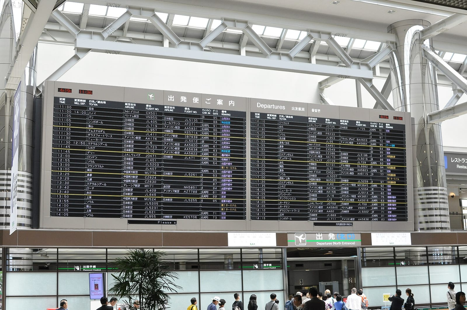 Narita Havalimanı 2. Terminal'de geniş bir kalkış panosu ve altında cam cepheli giriş bölümü, uzaktan görülen birkaç yolcu ile birlikte görülüyor.