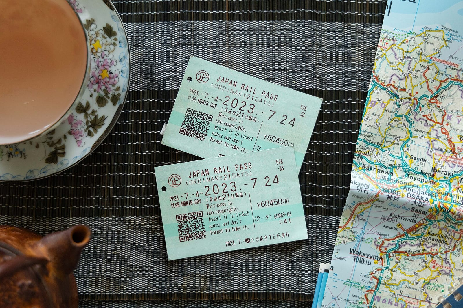 Ahşap bir masanın üzerinde iki Japan Rail Pass bileti, yanında bir çay fincanı, demlik ve Japonya haritası duruyor.