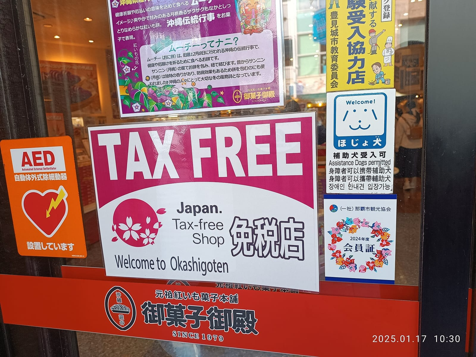 Naha'daki bir pastane mağazasının camına asılmış, pembe çiçek motifli ve Japonca ile İngilizce yazılı Tax Free vergi iadesi levhası yakından görünüyor.