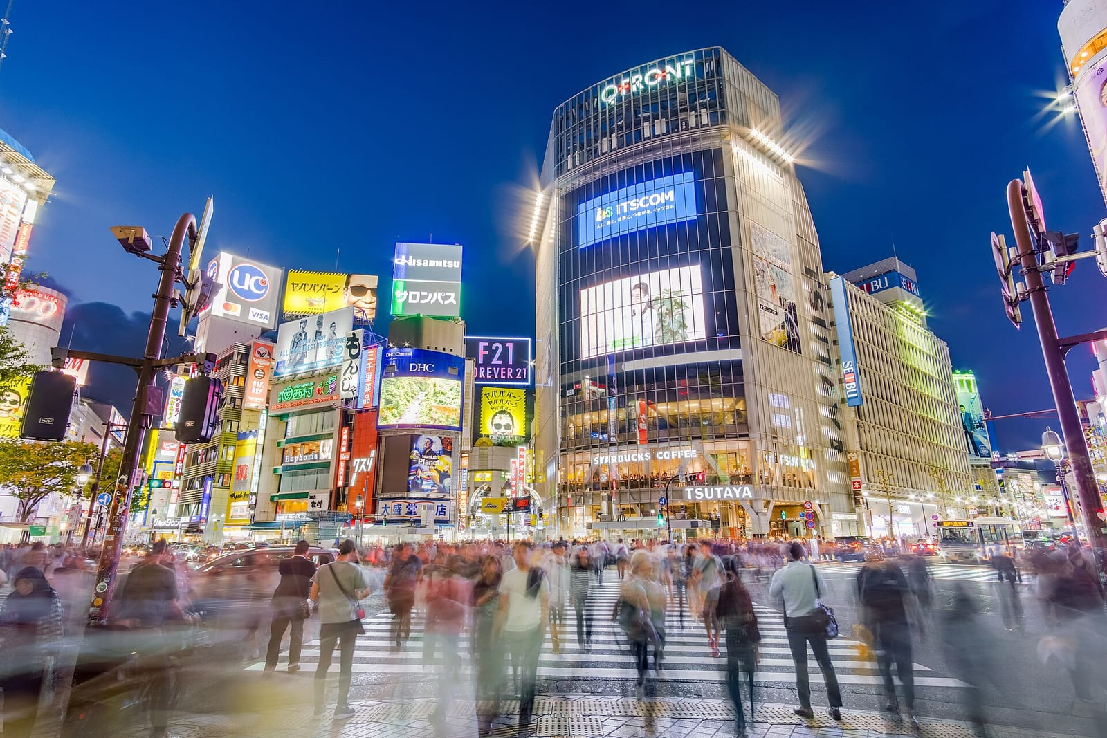 Akşam alacakaranlığında Shibuya Kavşağı'nda yürüyen kalabalık hareket bulanıklığıyla görünüyor, arkada parlak reklam panoları ve binalar yükseliyor.