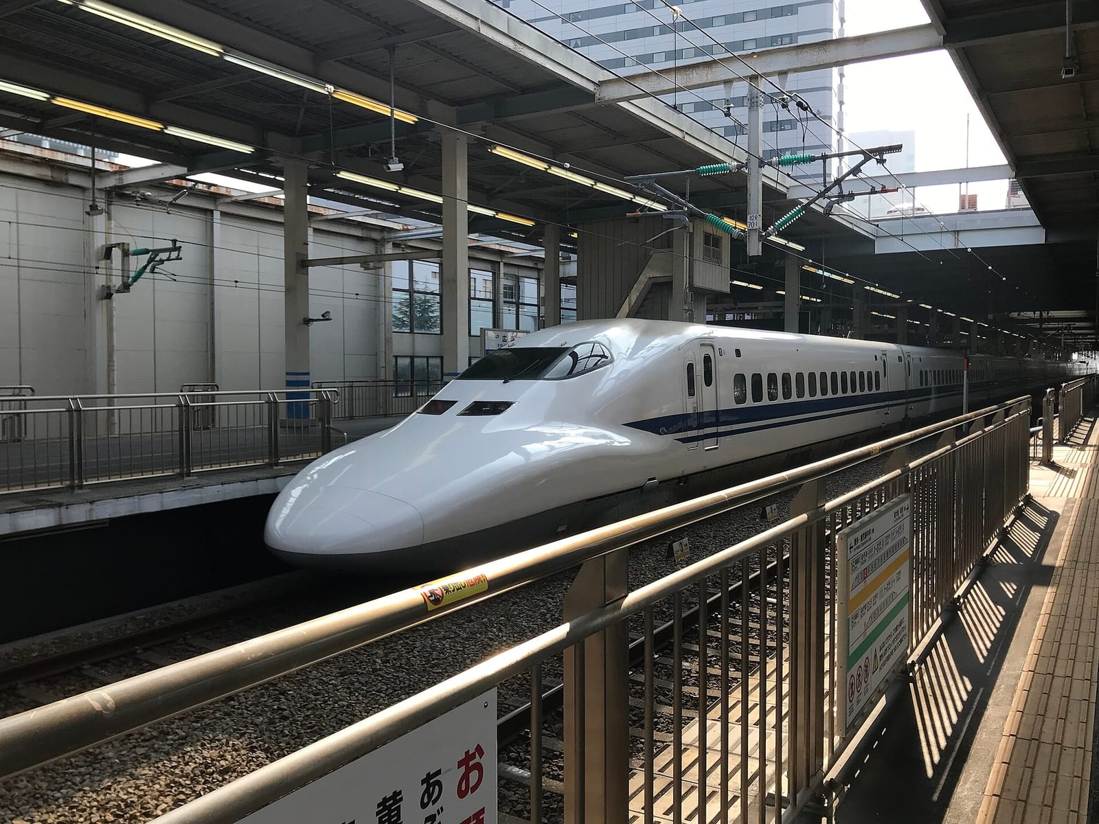 Hiroşima İstasyonu'nda peronda duran beyaz-mavi Sanyo Shinkansen treni, korkuluk ve üstü kapalı peron çatısıyla birlikte görülüyor.
