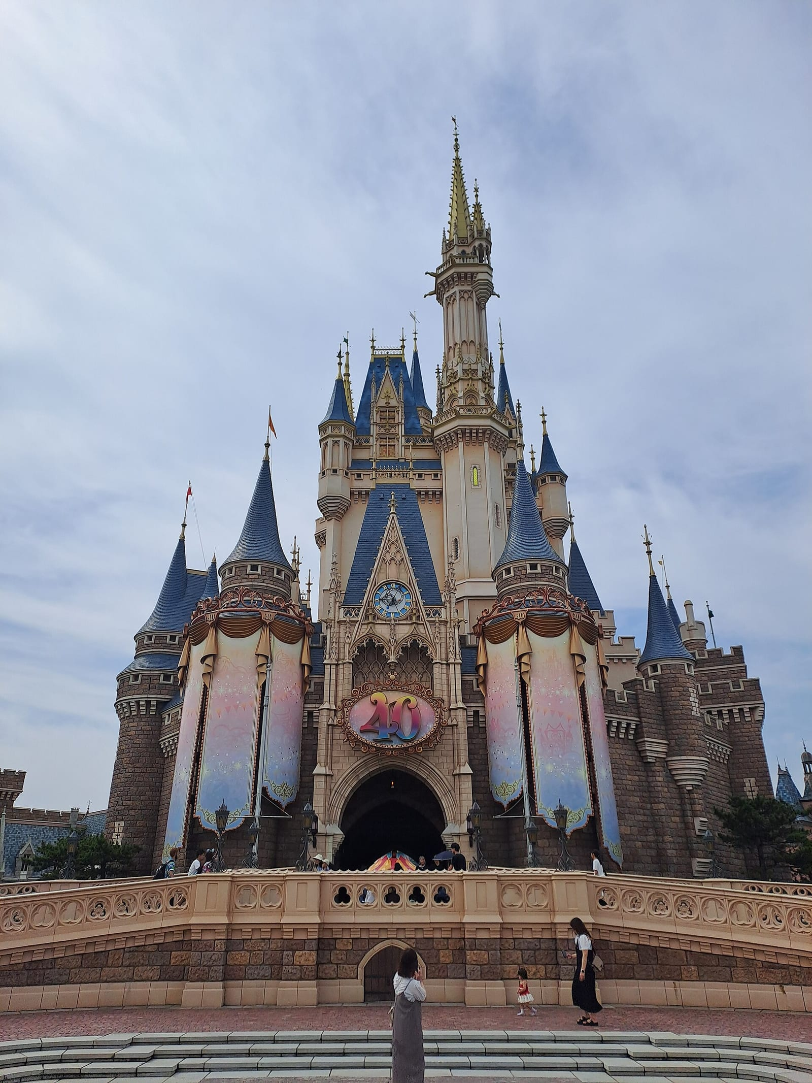 Tokyo Disneyland'deki Sinderella Şatosu, açık mavi gökyüzü altında önündeki merdivenler ve ziyaretçilerle birlikte gündüz çekilmiş.
