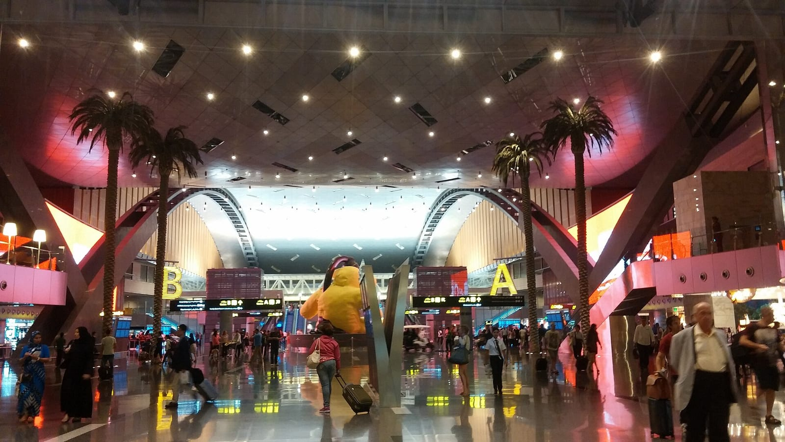 Doha Hamad Uluslararası Havalimanı'nın geniş terminal salonunda palmiye ağaçları, A ve B kapı tabelaları ile yürüyen yolcular görülüyor.