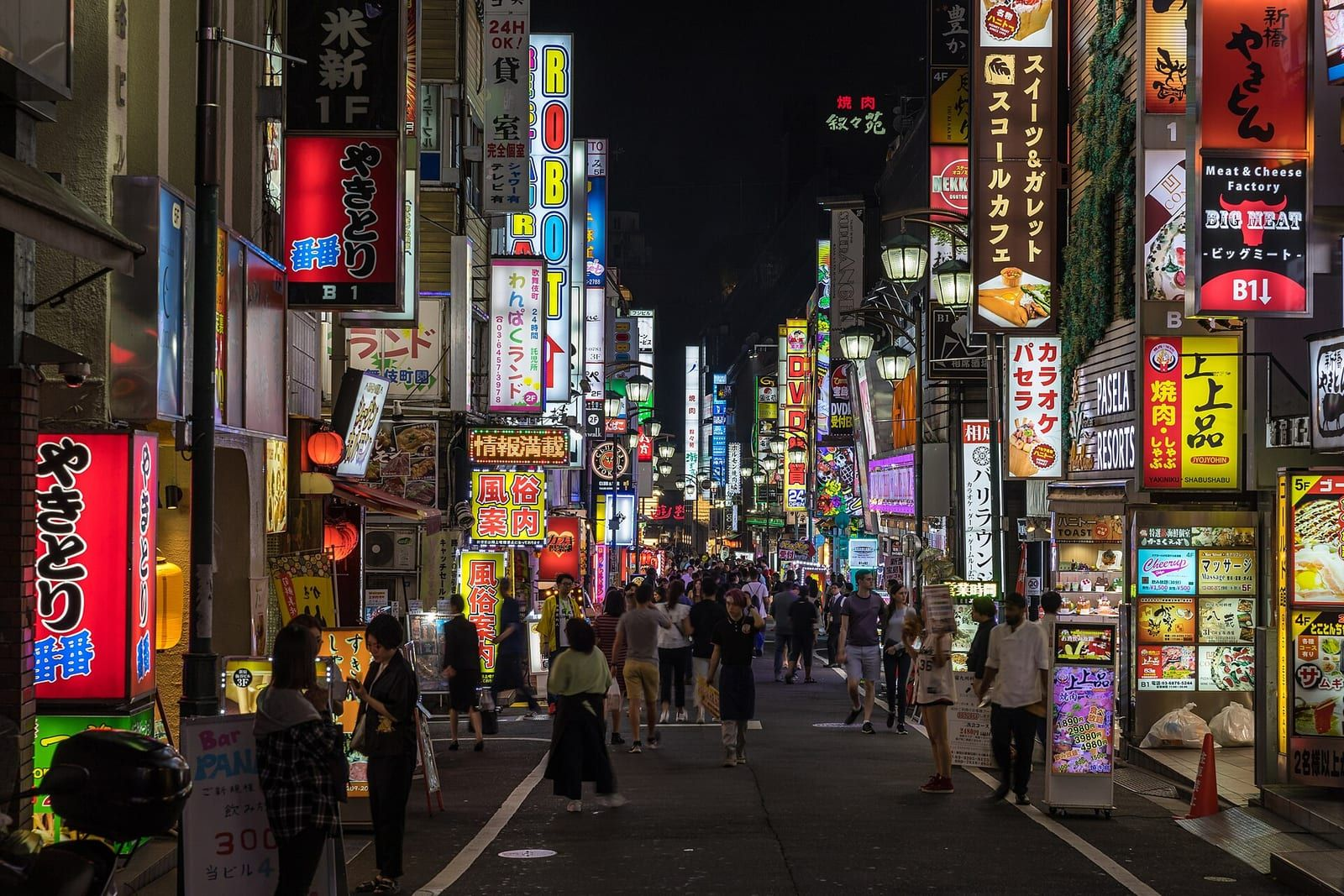 Tokyo Shinjuku Kabukichō'de gece neon tabelalarla dolu bir sokakta yürüyen insanlar ve iki yanda parlak renkli reklam panoları görülüyor.
